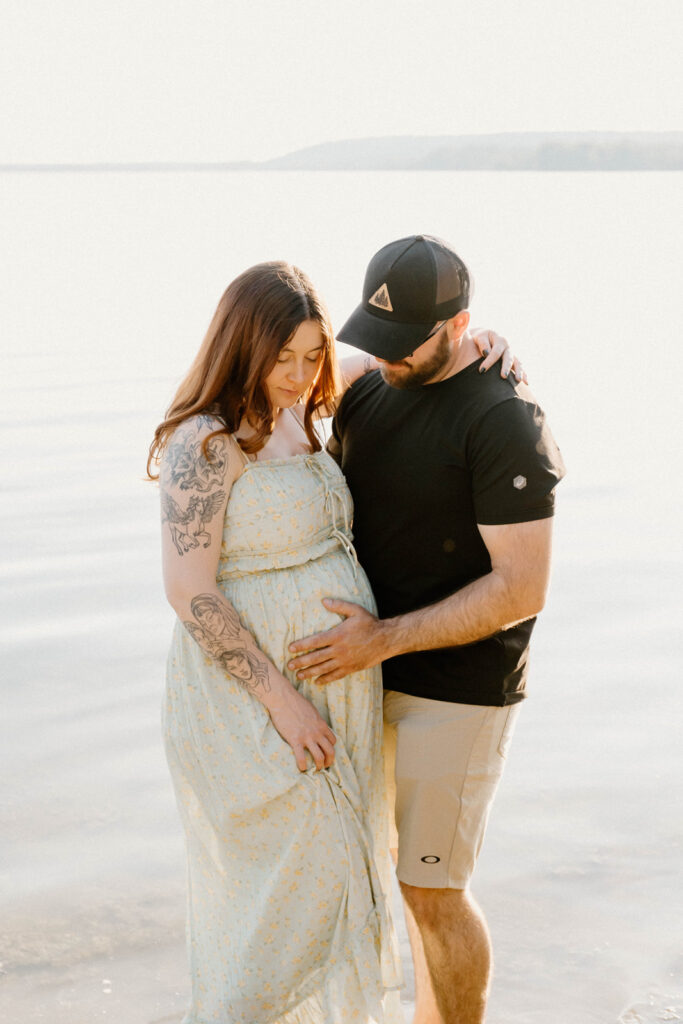 Couples maternity session at the beach in Round Lake in Eganville Ontario. The couple standing in the water with the sunset behind them