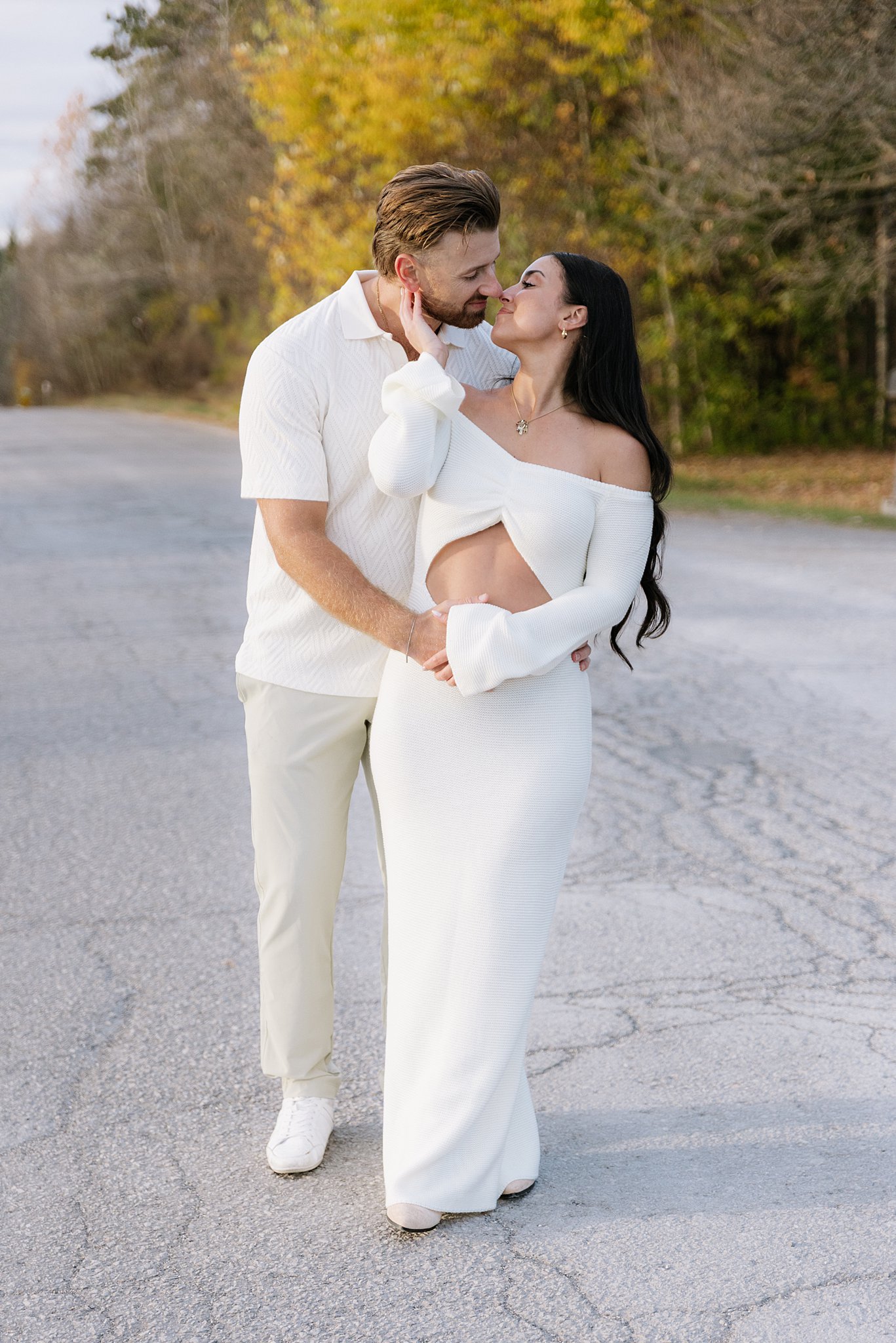Couple in white outfits sharing a kiss on a paved road with autumn-colored trees in the background.